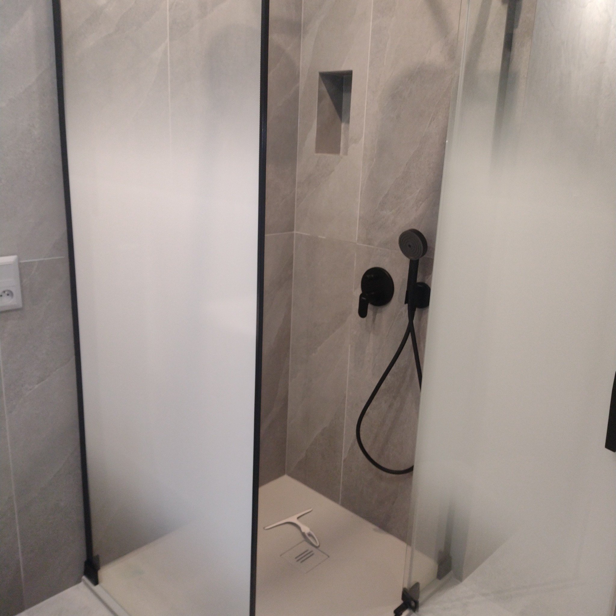 Nowoczesna kabina prysznicowa z szarymi płytkami imitującymi kamień, czarną armaturą i odpływem liniowym. W kabinie widoczna ściągaczka do wody.