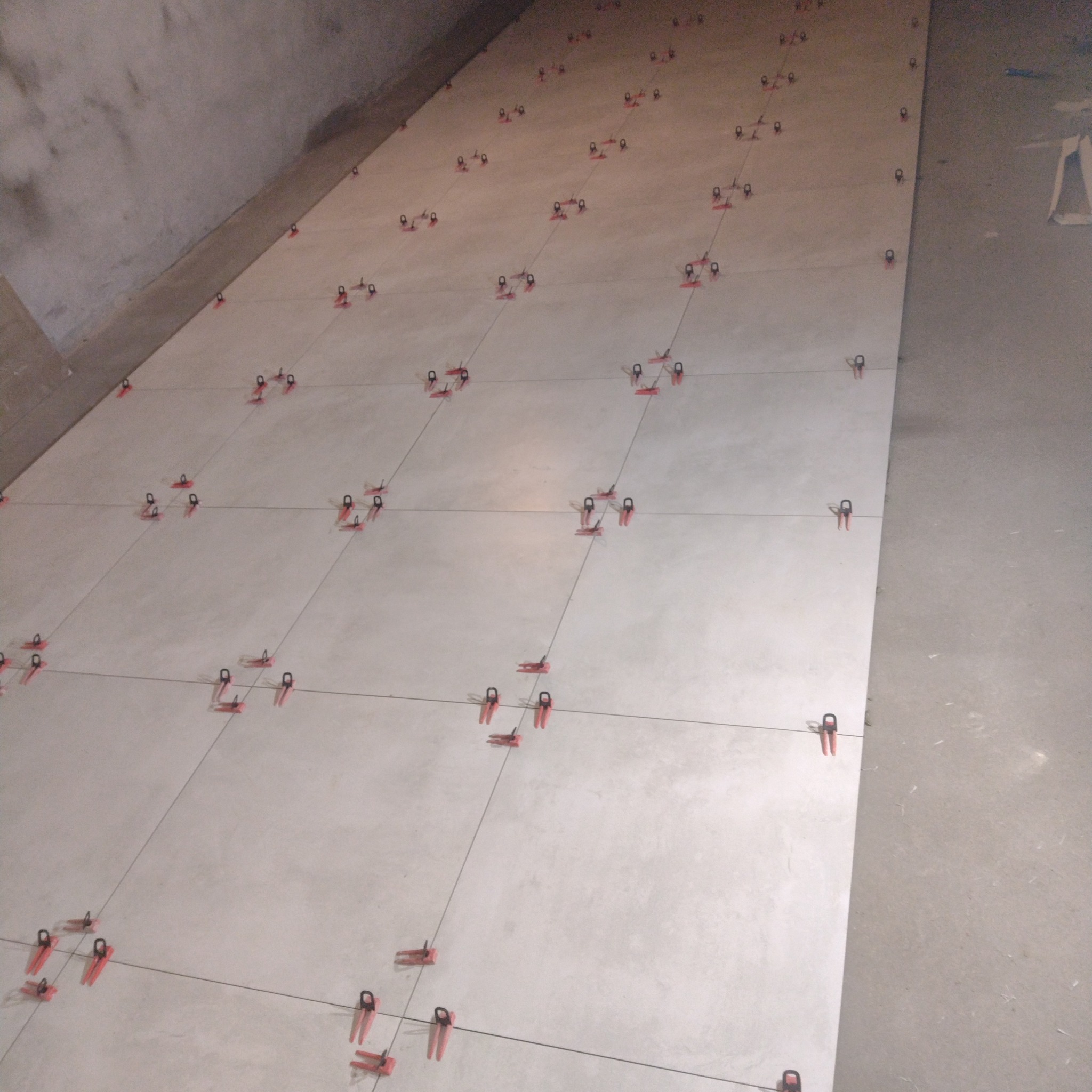 Układanie jasnych płytek podłogowych z użyciem czerwonych klipsów poziomujących i sznurka, widok z góry na niedokończoną powierzchnię.