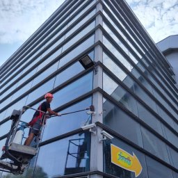 AlpinTop - Pracownik w czerwonym kasku na podnośniku koszowym myje szklaną elewację biurowca z licznymi kamerami monitoringu.