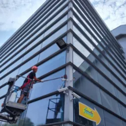 Pracownik w czerwonym kasku na podnośniku koszowym myje szklaną elewację biurowca z licznymi kamerami monitoringu.