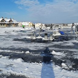 AlpinTop - Pracownicy z łopatami usuwają śnieg i lód z płaskiego dachu budynku w słoneczny dzień. W tle widoczne domy jednorodzinne i błękitne niebo z chmurami.