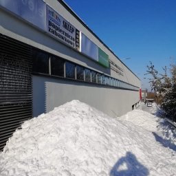 AlpinTop - Ogromna pryzma śniegu usunięta z dachu budynku usługowego z widocznymi szyldami sklepów, cień osoby robiącej zdjęcie na śniegu, błękitne niebo w tle.