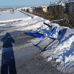 AlpinTop - Dwie niebieskie łopaty do śniegu leżą na płaskim dachu pokrytym śniegiem i lodem w słoneczny dzień, w tle budynki mieszkalne i cień osoby robiącej zdjęcie.
