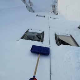 AlpinTop - Niebieska łopata do śniegu leży na dachu pokrytym grubą warstwą śniegu, widoczne okna dachowe i fragment ogrodu w tle.