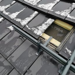 AlpinTop - Fragment dachu pokrytego czarną dachówką, częściowo pokrytą śniegiem, z widocznym fragmentem konstrukcji dachu i izolacji, z zamontowanymi metalowymi elementami zabezpieczającymi.