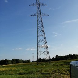 AlpinTop - Wysoka, stalowa wieża energetyczna na tle błękitnego nieba i zielonego pola, dwie osoby pracują na wysokości, w tle widoczna kolejna wieża i las.