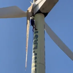 Pracownik w uprzęży na turbinie wiatrowej z uszkodzonymi łopatami, widok z dołu na tle błękitnego nieba, napis GREPLAST na wieży.