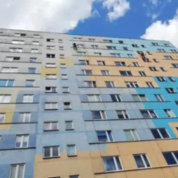 Wysoki blok mieszkalny w trakcie malowania elewacji; dwóch pracowników na linach alpinistycznych nanosi farbę na fragmenty budynku, widoczne niepomalowane szare partie oraz pomalowane na niebiesko...