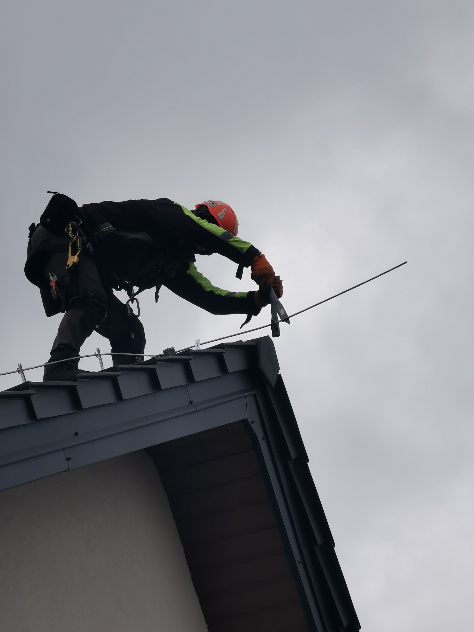 Pracownik w kasku i uprzęży asekuracyjnej, pochylony nad krawędzią dachu z szarej blachy, trzyma w rękach narzędzie, widoczne stalowe linki zabezpieczające.