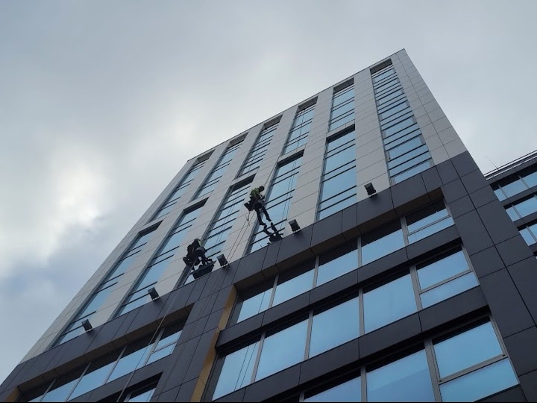 Dwóch alpinistów przemysłowych na linach podczas pracy na elewacji wysokiego biurowca z szarymi panelami i niebieskimi szybami, widok z dołu pod kątem w pochmurny dzień.