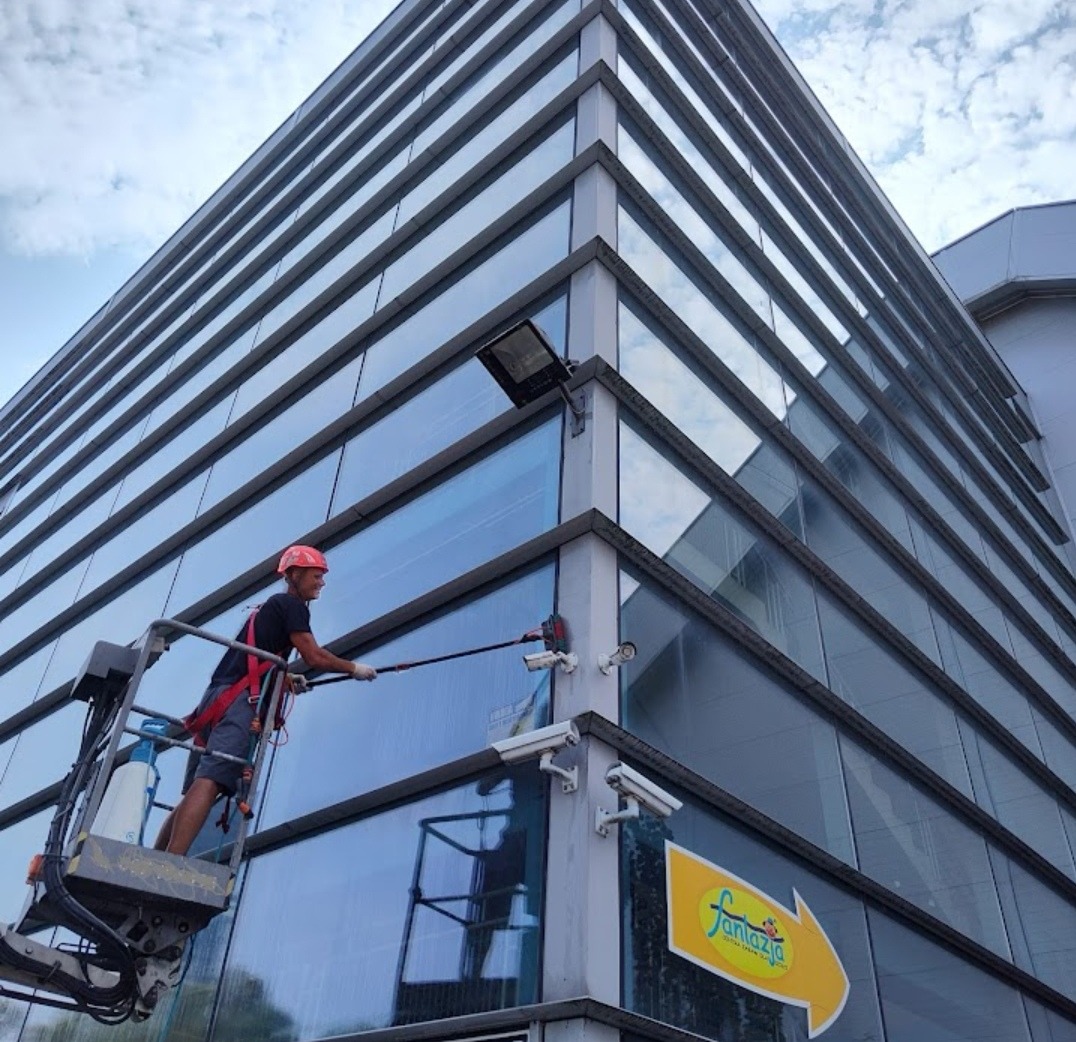 Pracownik w czerwonym kasku na podnośniku koszowym myje szklaną elewację biurowca z licznymi kamerami monitoringu.