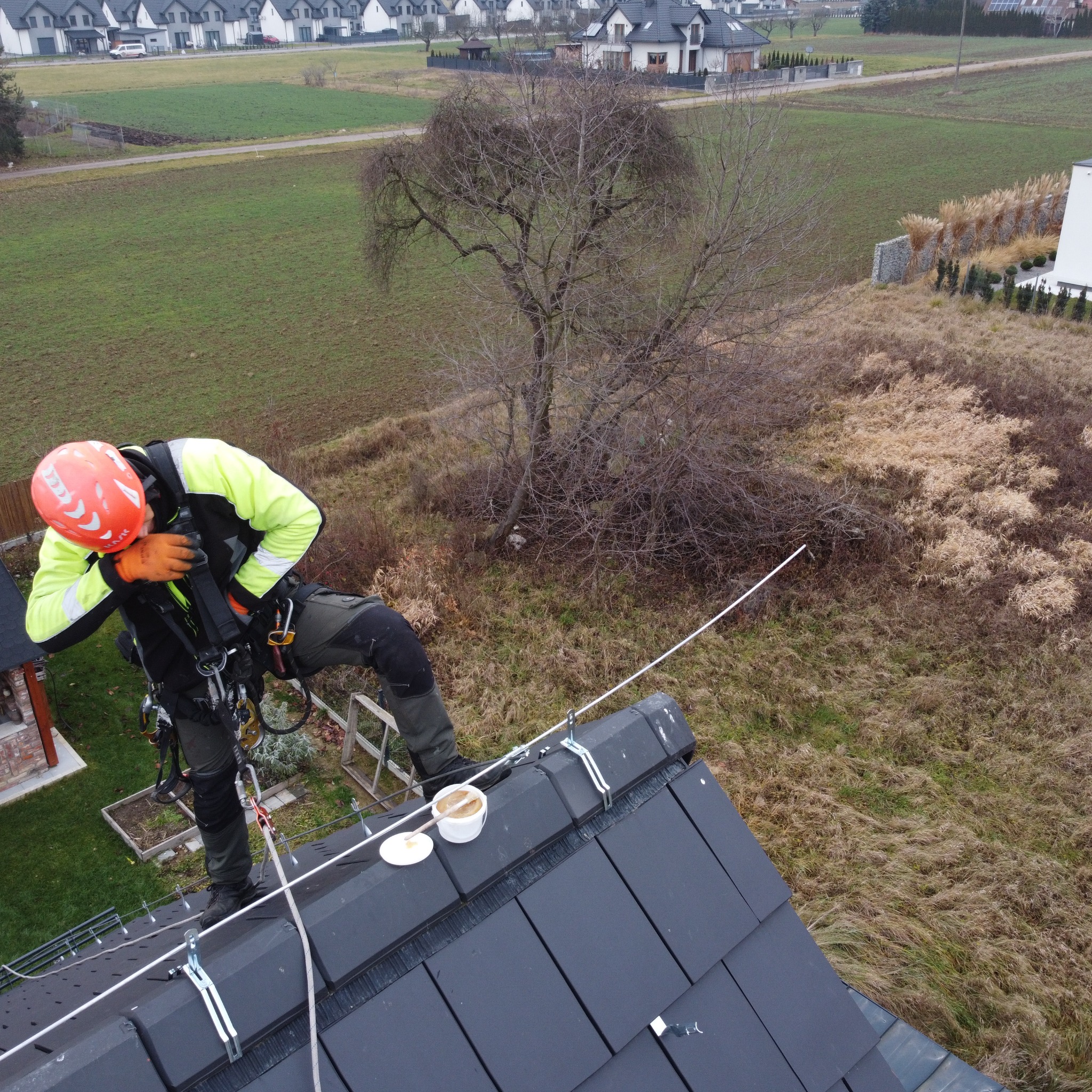 Pracownik w kasku i uprzęży na stromym dachu z czarnej dachówki, zabezpieczony linami, z widocznym pojemnikiem z masą uszczelniającą i pędzlem, w tle zabudowa jednorodzinna i zieleń.