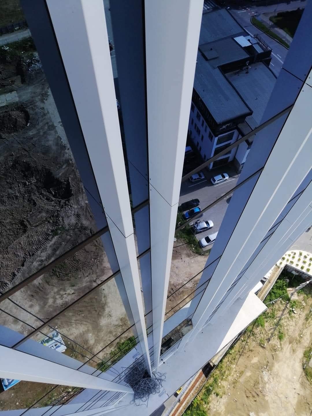 Widok z góry na fragment szklanej elewacji budynku w trakcie prac, widoczne liny zabezpieczające i otoczenie budynku z placem budowy i parkingiem.