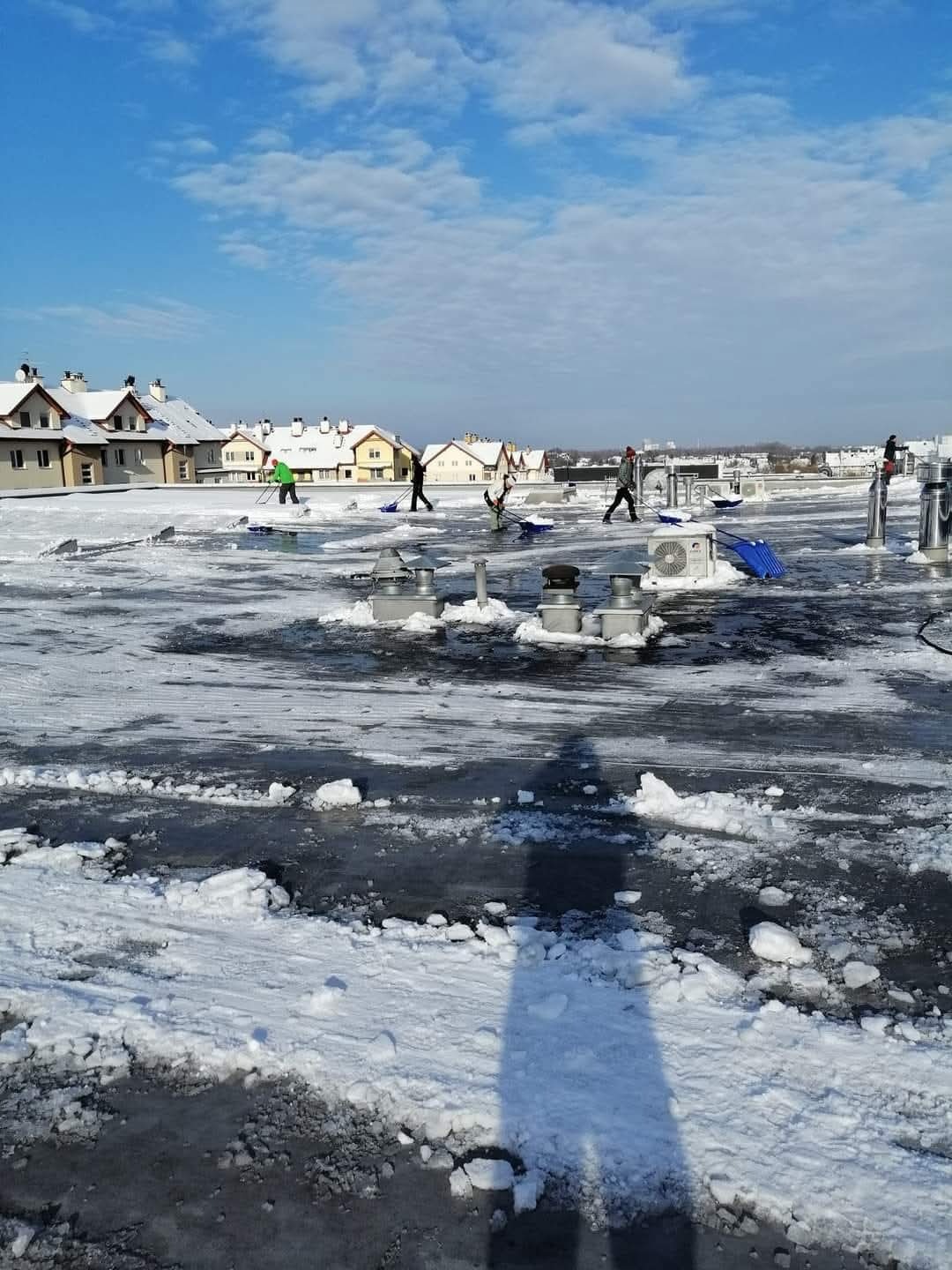Pracownicy z łopatami usuwają śnieg i lód z płaskiego dachu budynku w słoneczny dzień. W tle widoczne domy jednorodzinne i błękitne niebo z chmurami.
