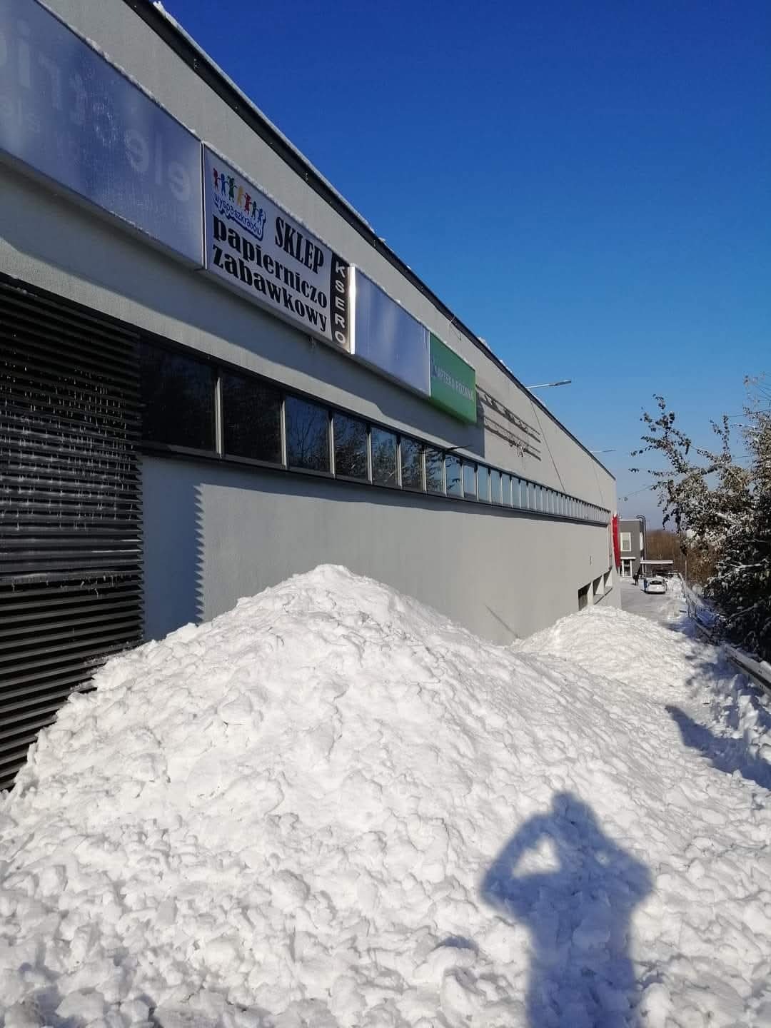 Ogromna pryzma śniegu usunięta z dachu budynku usługowego z widocznymi szyldami sklepów, cień osoby robiącej zdjęcie na śniegu, błękitne niebo w tle.
