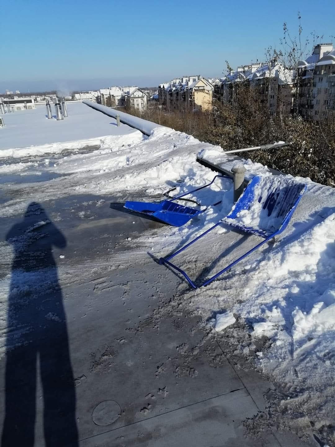 Dwie niebieskie łopaty do śniegu leżą na płaskim dachu pokrytym śniegiem i lodem w słoneczny dzień, w tle budynki mieszkalne i cień osoby robiącej zdjęcie.