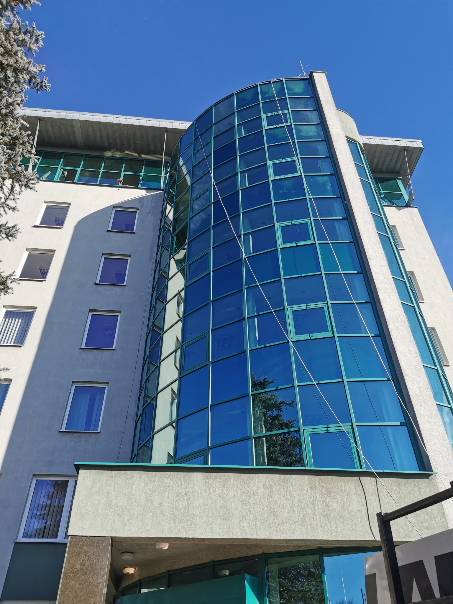 Wysoki budynek biurowy z przeszkloną, zaokrągloną fasadą, widziany z dołu pod kątem, z widocznymi linami zabezpieczającymi na szklanej powierzchni, sugerującymi prace na wysokości.