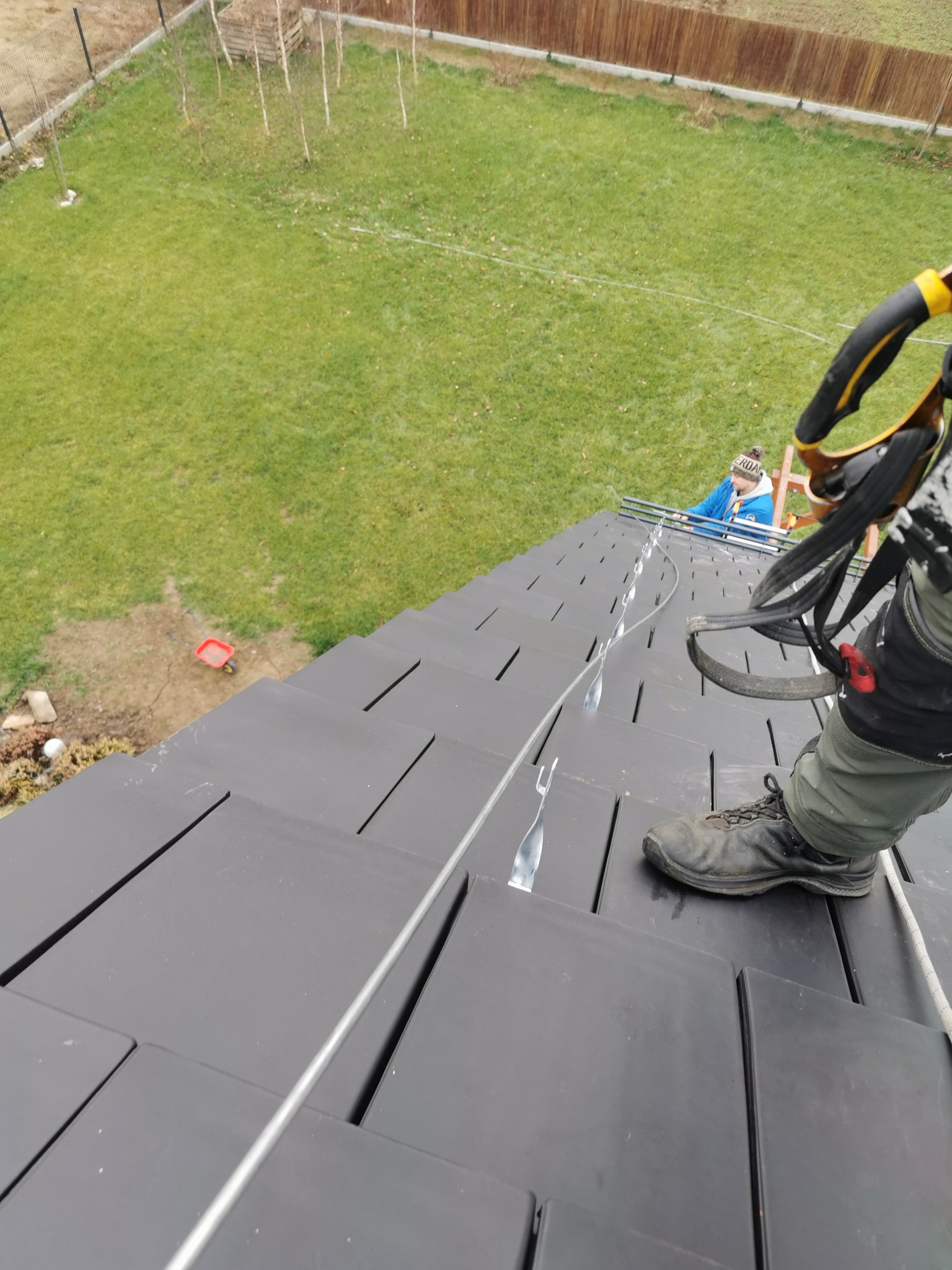 Pracownik w uprzęży zabezpieczającej stoi na ciemnym dachu z płyt, widoczna lina asekuracyjna i drugi pracownik w tle, trawnik i ogrodzenie.