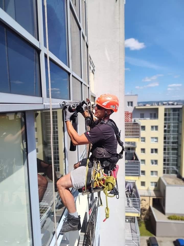 Pracownik w kasku i uprzęży, zawieszony na linie, wierci otwór w ramie okiennej wysokiego budynku; w tle zabudowa miejska.