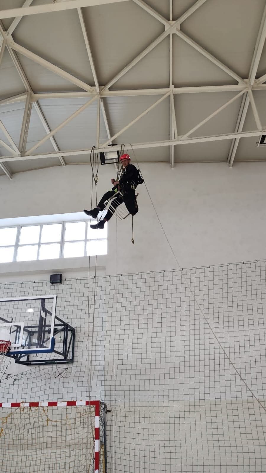 Pracownik w czerwonym kasku, ubrany w kombinezon, zawieszony na linach i platformie, wykonuje prace wysokościowe wewnątrz hali sportowej z siatką zabezpieczającą i koszem do koszykówki w tle.