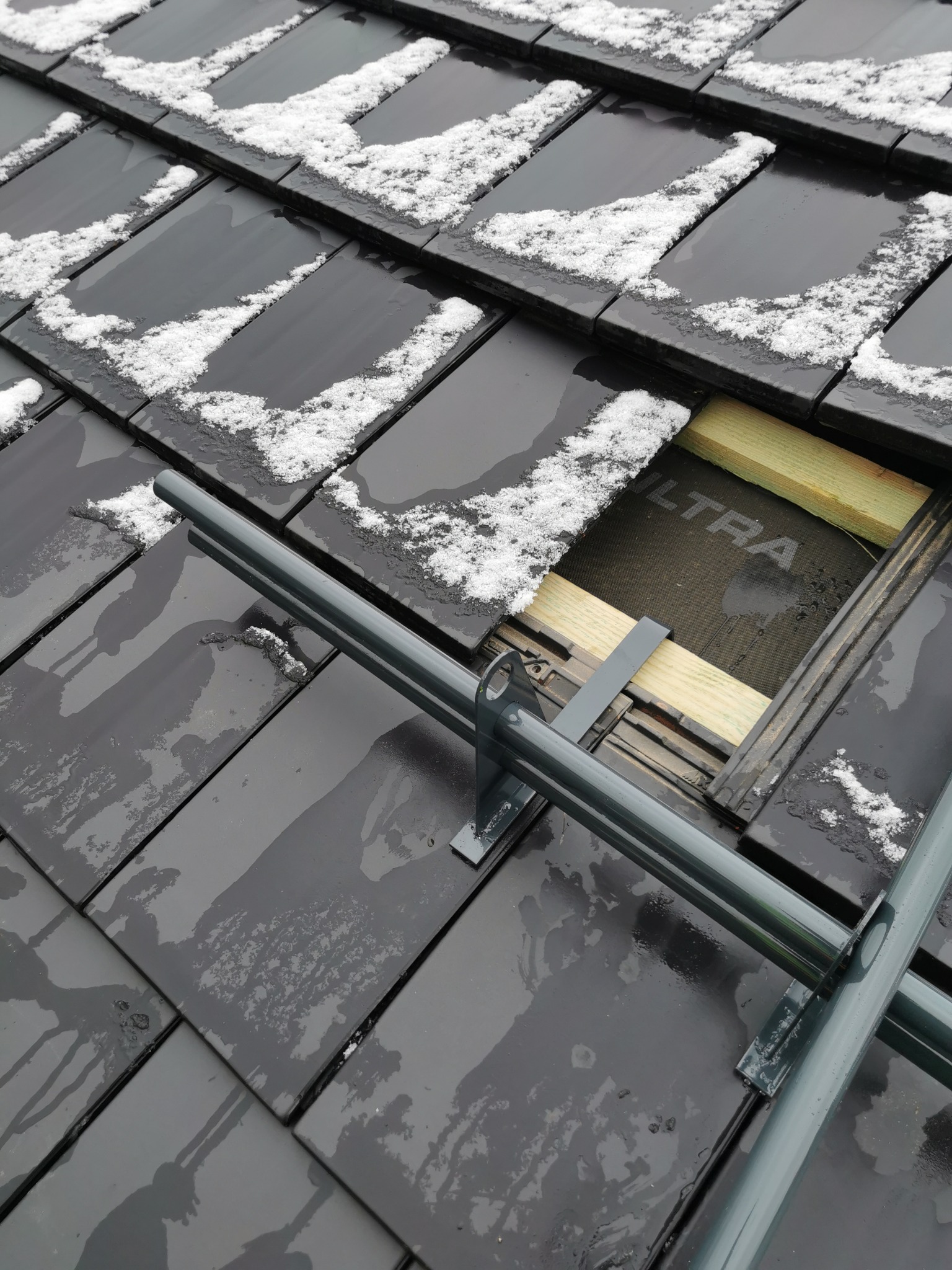 Fragment dachu pokrytego czarną dachówką, częściowo pokrytą śniegiem, z widocznym fragmentem konstrukcji dachu i izolacji, z zamontowanymi metalowymi elementami zabezpieczającymi.