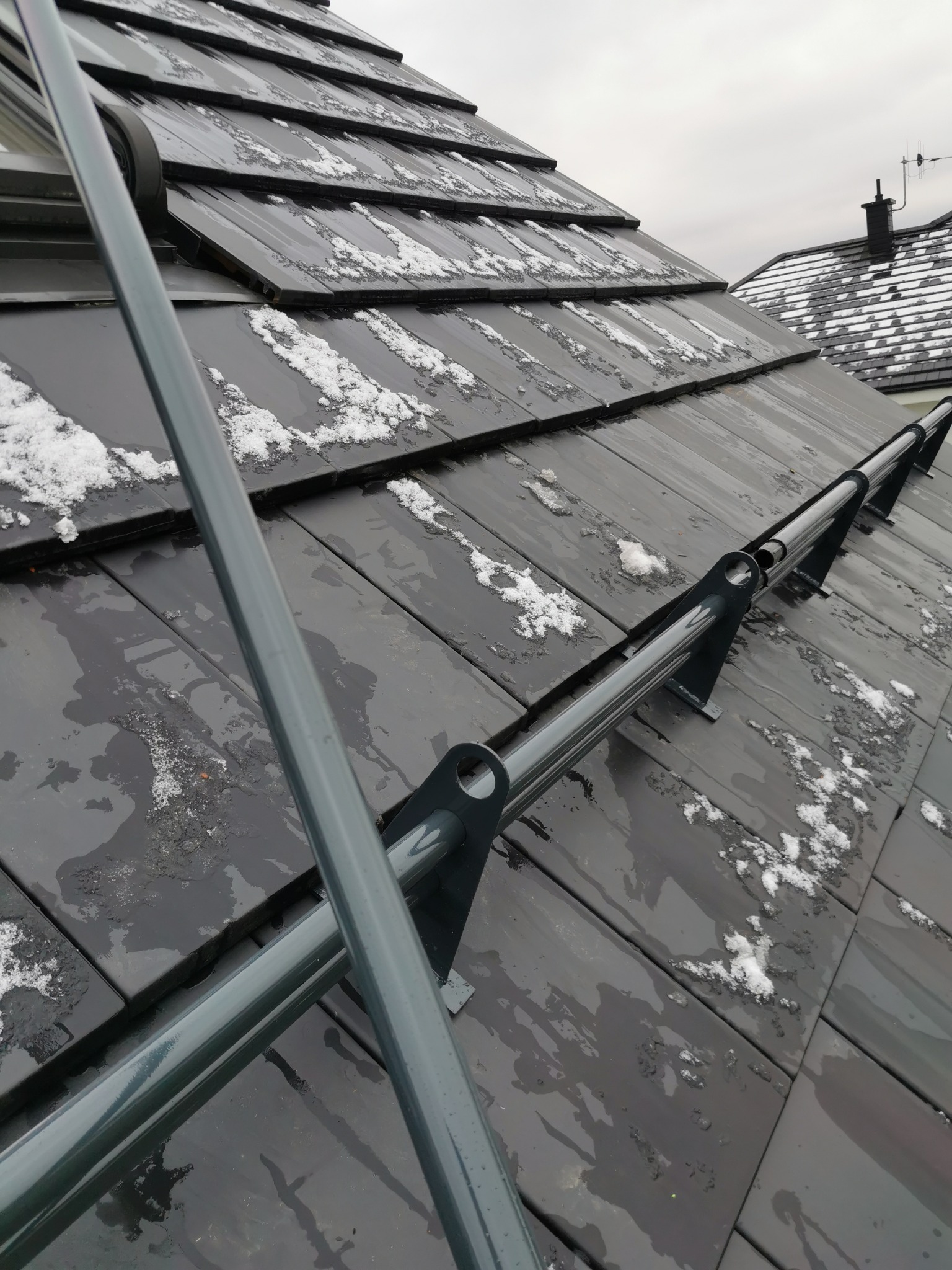 Ciemna, mokra dachówka z resztkami śniegu i metalowymi elementami systemu zabezpieczającego przed upadkiem z wysokości.
