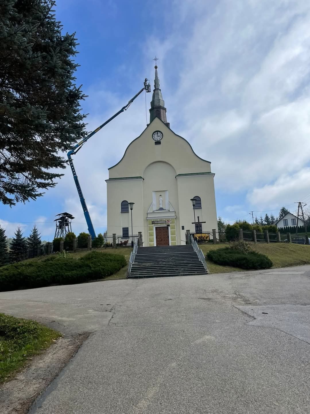 Pracownik na podnośniku koszowym przy szczycie kościoła, prawdopodobnie podczas prac konserwacyjnych lub naprawczych, widok z dołu na budynek i podnośnik na tle pochmurnego nieba.