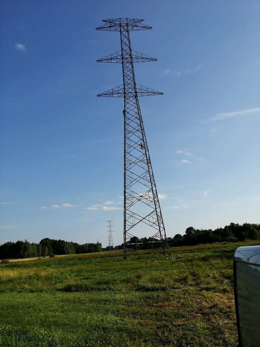Wysoka, stalowa wieża energetyczna na tle błękitnego nieba i zielonego pola, dwie osoby pracują na wysokości, w tle widoczna kolejna wieża i las.