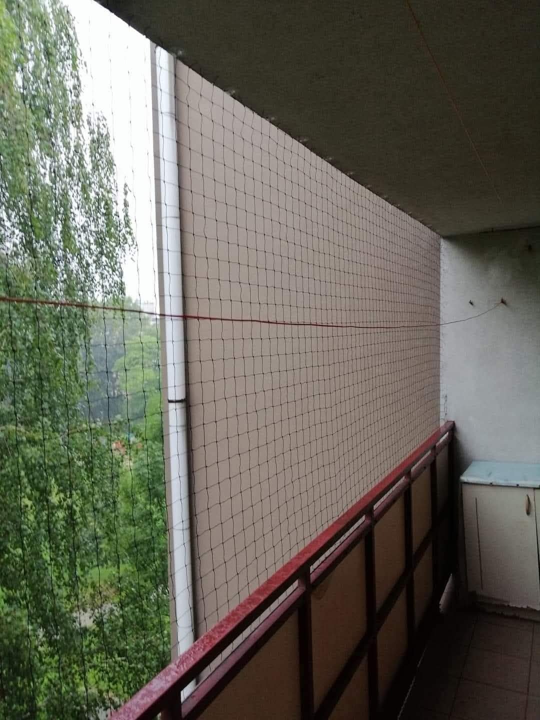 Zabezpieczony siatką balkon w bloku, widok na zieleń za siatką, czerwona poręcz, biała rynna na ścianie.