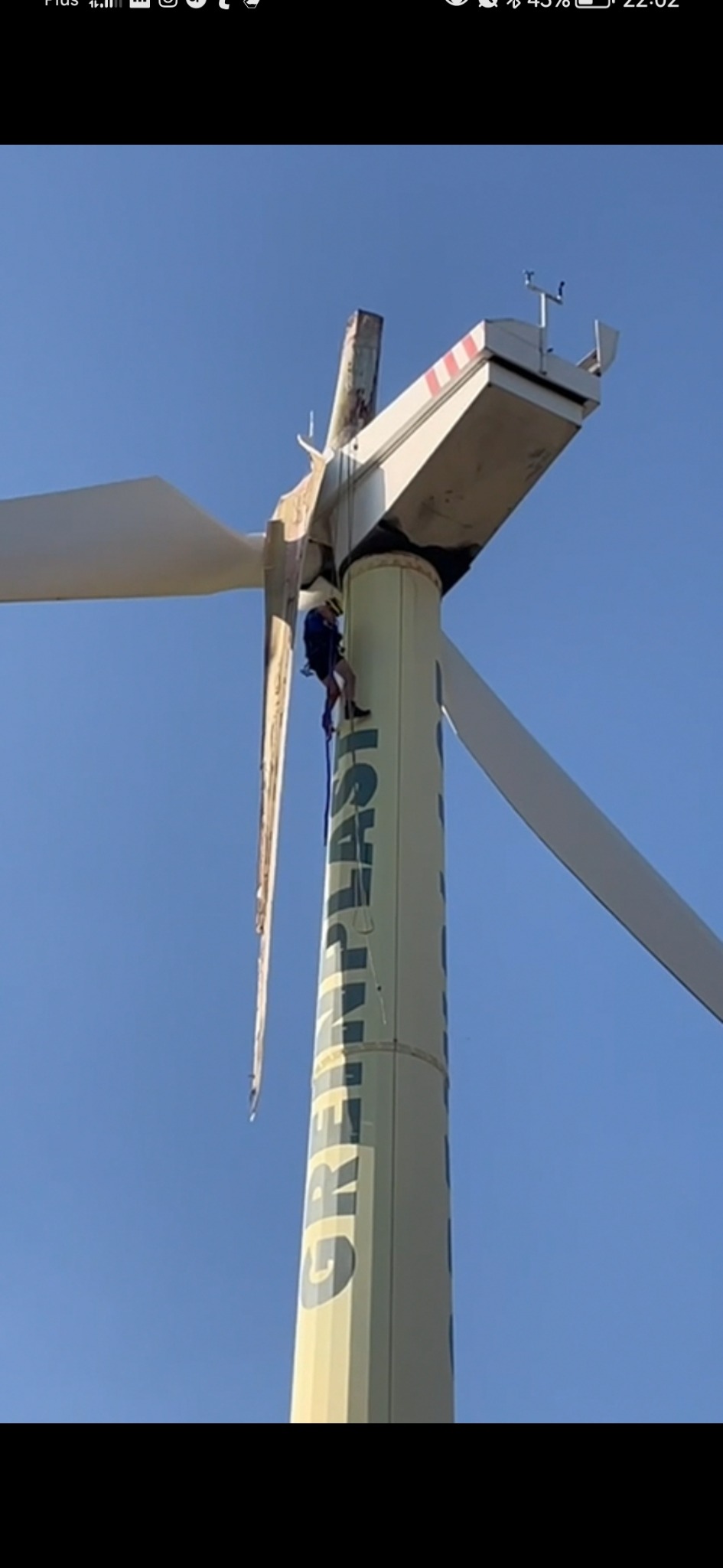 Pracownik w uprzęży na turbinie wiatrowej z uszkodzonymi łopatami, widok z dołu na tle błękitnego nieba, napis GREPLAST na wieży.