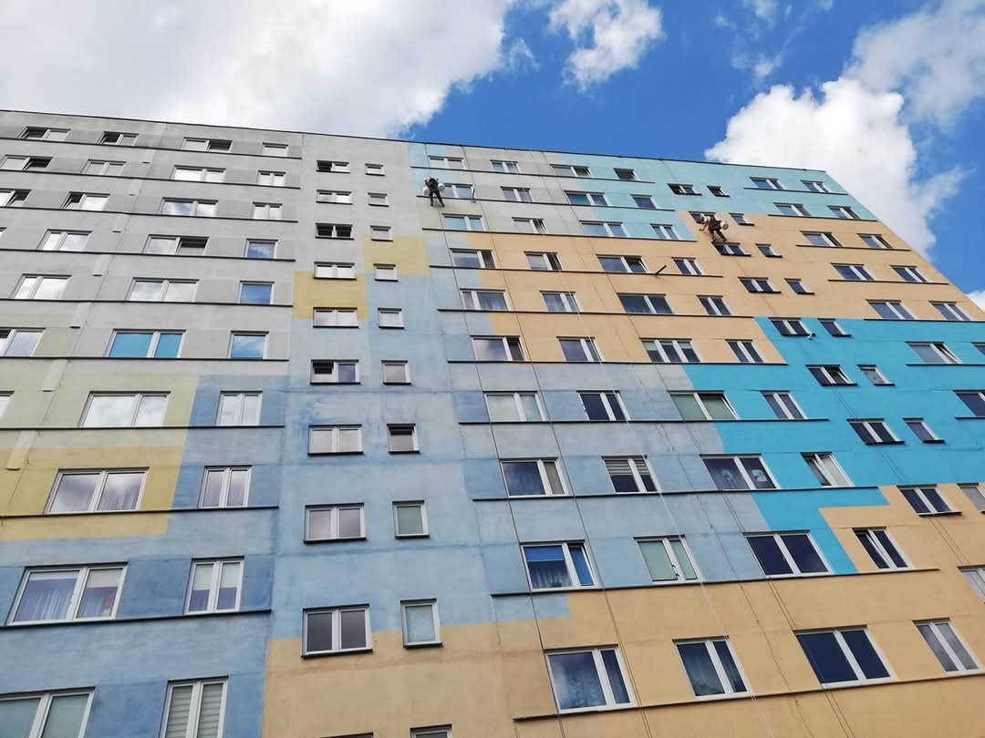 Wysoki blok mieszkalny w trakcie malowania elewacji; dwóch pracowników na linach alpinistycznych nanosi farbę na fragmenty budynku, widoczne niepomalowane szare partie oraz pomalowane na niebiesko...