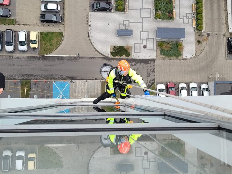 Pracownik w kasku i odblaskowej kurtce, zabezpieczony linami, czyści szklaną elewację wysokiego budynku z perspektywy dachu, widok z góry na parking i otoczenie.