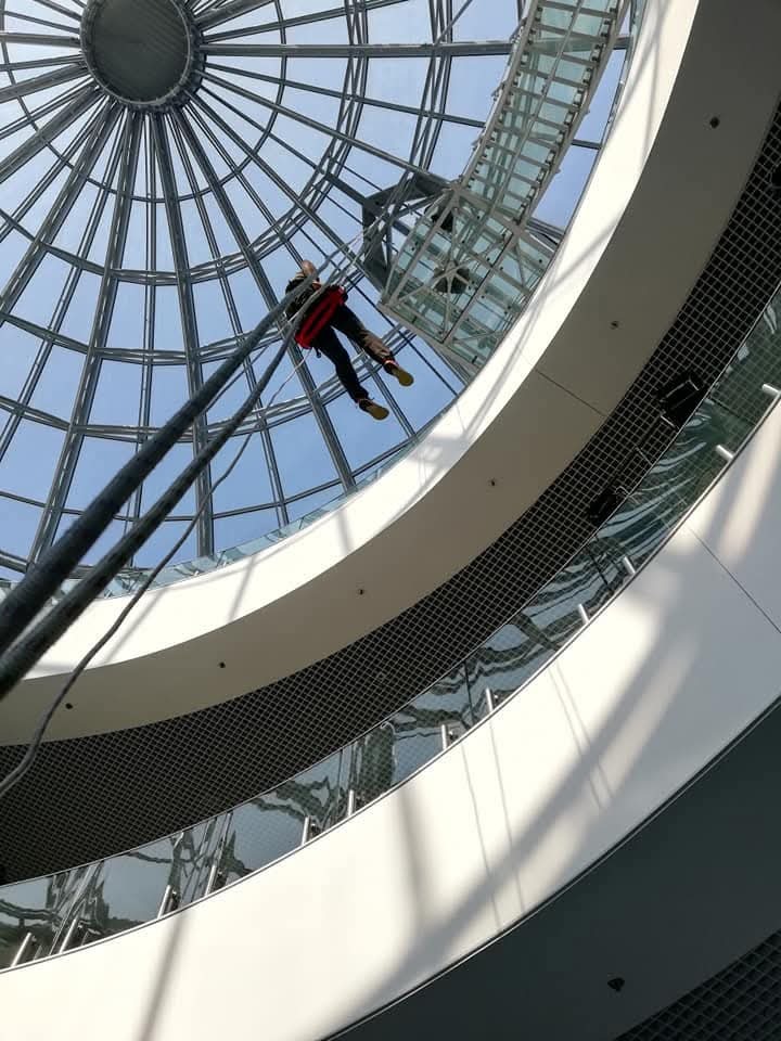 Pracownik na linach zabezpieczających, w trakcie pracy pod szklaną kopułą budynku, widok z dołu na konstrukcję i osobę w uprzęży.