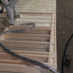 BEKO ARTS izolacje natryskowe pianą pur - Pracownik w kombinezonie aplikuje pianę izolacyjną w drewnianą konstrukcję szkieletową w hali, widoczny wąż urządzenia do natrysku, częściowo wypełnione przestrzenie między belkami.