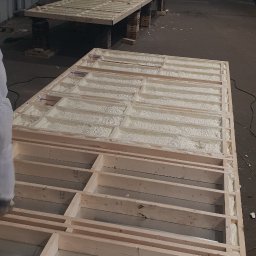 BEKO ARTS izolacje natryskowe pianą pur - Ocieplanie pianką PUR w drewnianej konstrukcji szkieletowej, widoczna metalowa płyta i wąż w folii, w tle konstrukcja na paletach w hali.
