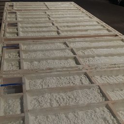 BEKO ARTS izolacje natryskowe pianą pur - Drewniana konstrukcja wypełniona białą pianką izolacyjną w hali, widok z góry, fragment niebieskiej taśmy na jednej z belek.