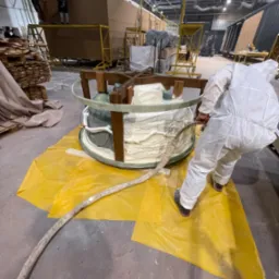 Pracownik w kombinezonie ochronnym aplikuje białą pianę izolacyjną do okrągłej formy z drewnianym stelażem na żółtej folii ochronnej w hali produkcyjnej.