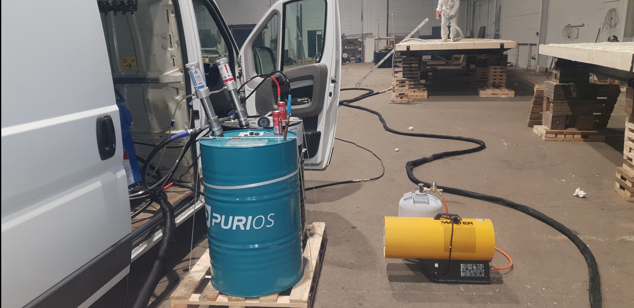 Sprzęt do natrysku pianki PUR: niebieska beczka z napisem PURIO, pompy dozujące, nagrzewnica MASTER i biały bus z otwartymi drzwiami w hali produkcyjnej, w tle pracownik w kombinezonie ochronnym...