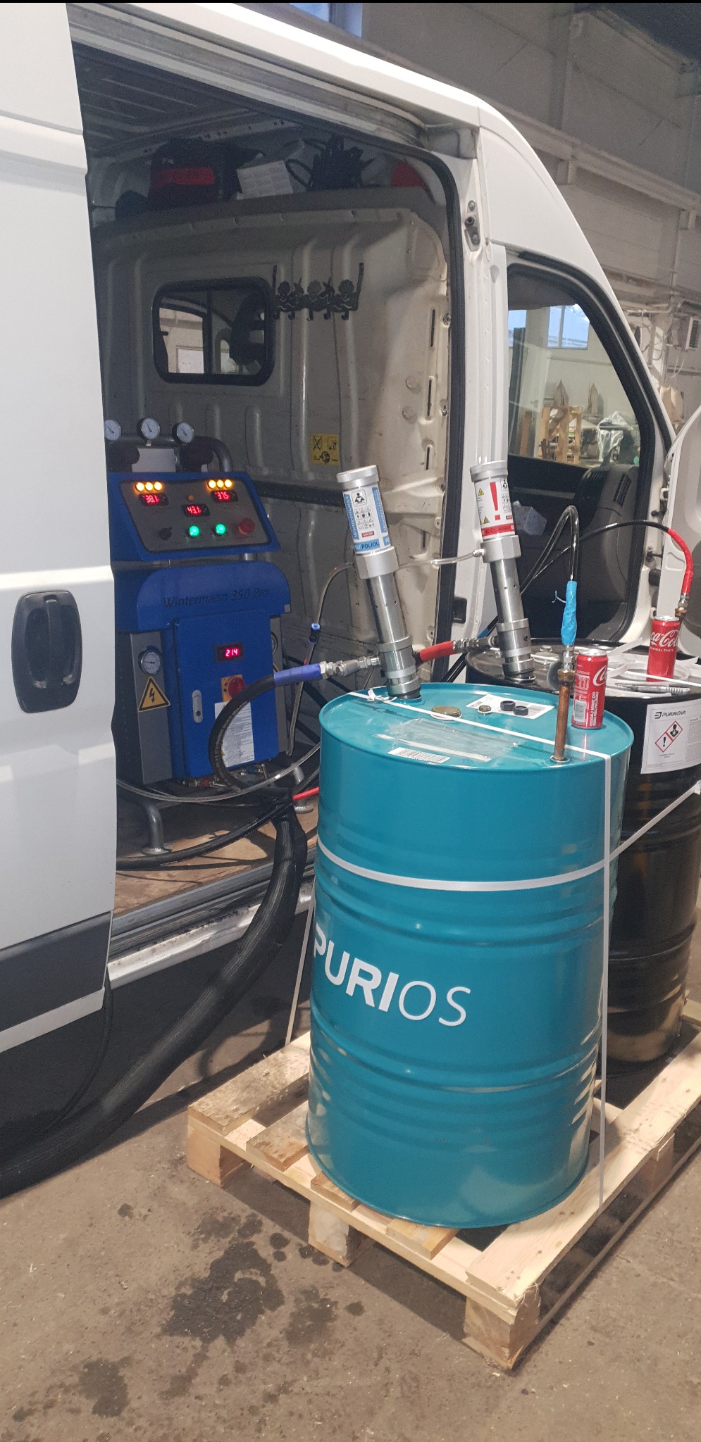 Sprzęt do natrysku pianki izolacyjnej PUR wewnątrz furgonetki: niebieski agregat Wintermann 350 Pro z wyświetlaczami, beczka z napisem PURiOS na palecie, dwa pojemniki z poliolami, puszki Coca-Coli...