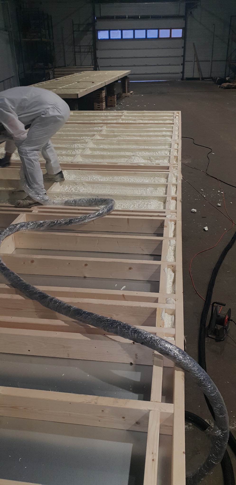 Pracownik w kombinezonie aplikuje pianę izolacyjną w drewnianą konstrukcję szkieletową w hali, widoczny wąż urządzenia do natrysku, częściowo wypełnione przestrzenie między belkami.