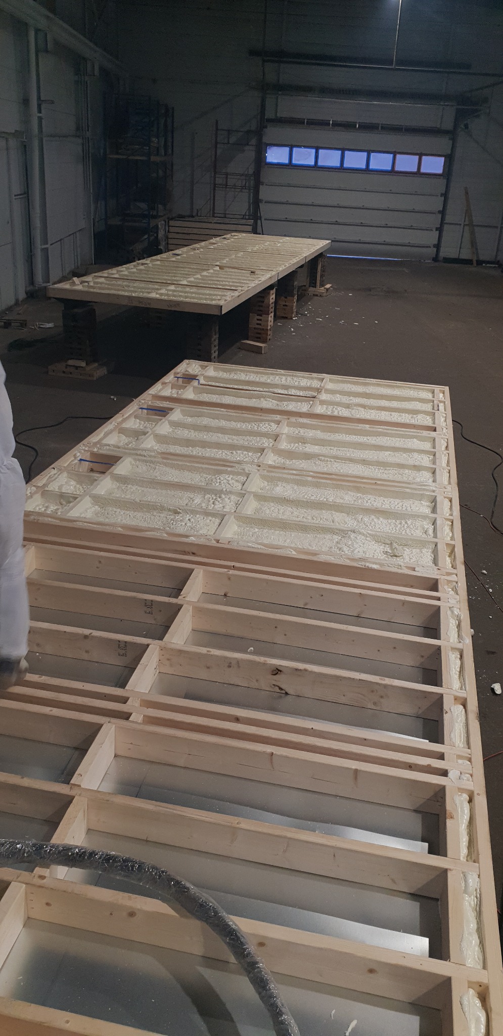 Ocieplanie pianką PUR w drewnianej konstrukcji szkieletowej, widoczna metalowa płyta i wąż w folii, w tle konstrukcja na paletach w hali.