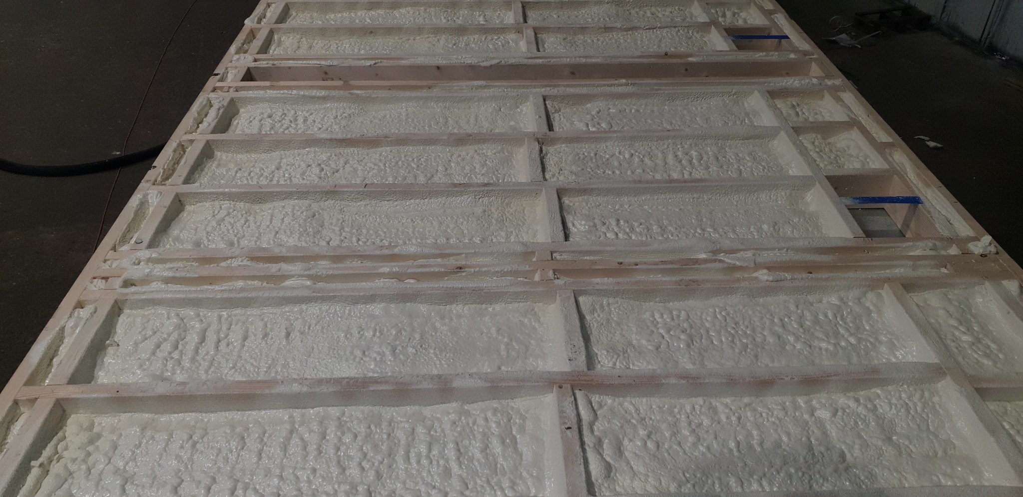 Drewniana konstrukcja wypełniona białą pianą izolacyjną, widok z góry, częściowo widoczny przewód doprowadzający pianę.