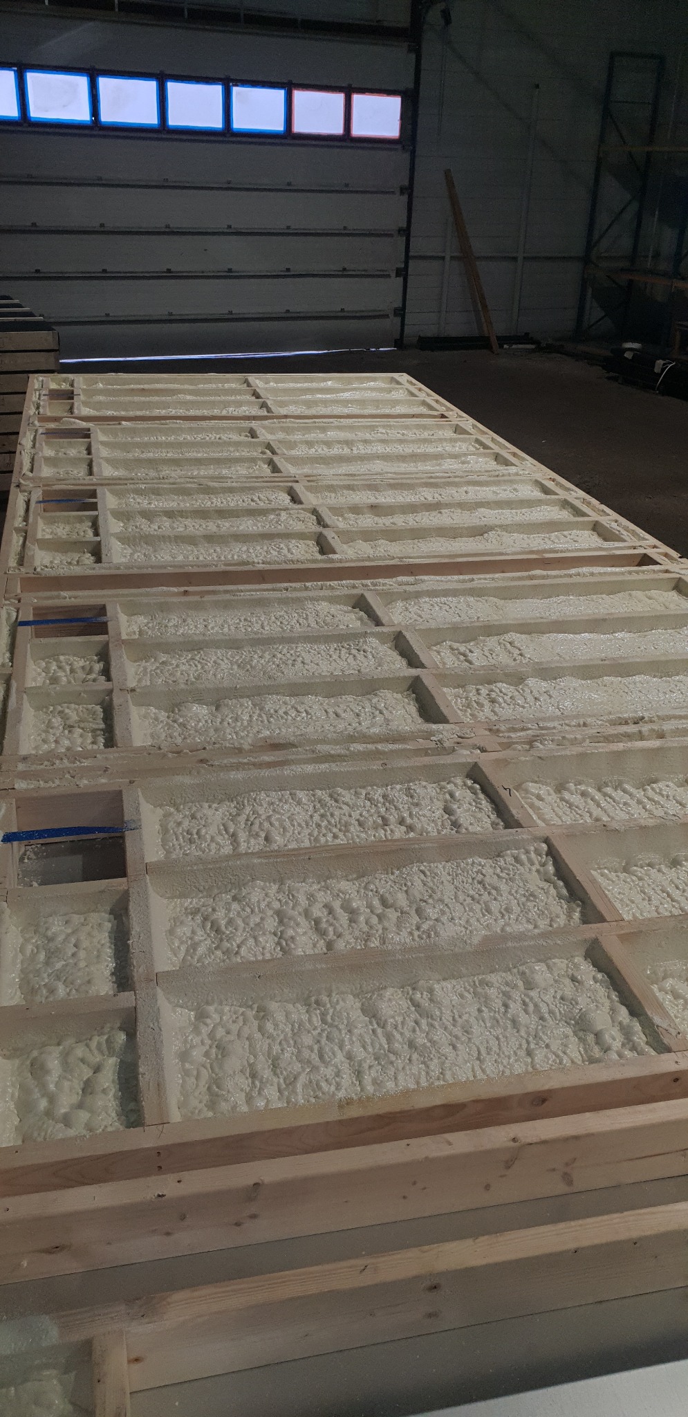 Drewniana konstrukcja wypełniona białą pianką izolacyjną w hali, widok z góry, fragment niebieskiej taśmy na jednej z belek.