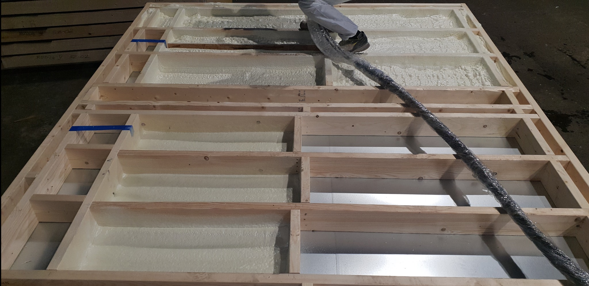 Drewniana konstrukcja szkieletowa wypełniona jasną pianą izolacyjną, widoczny czarny wąż doprowadzający pianę i fragment metalowej płyty w dolnej części.