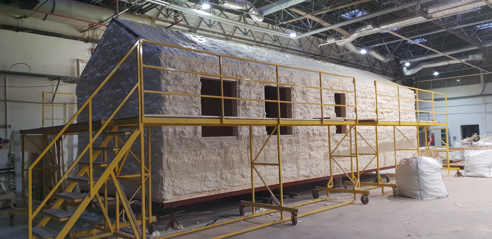Modułowy dom w hali produkcyjnej, pokryty pianką izolacyjną, z żółtym rusztowaniem, widoczne otwory okienne i elementy konstrukcyjne.