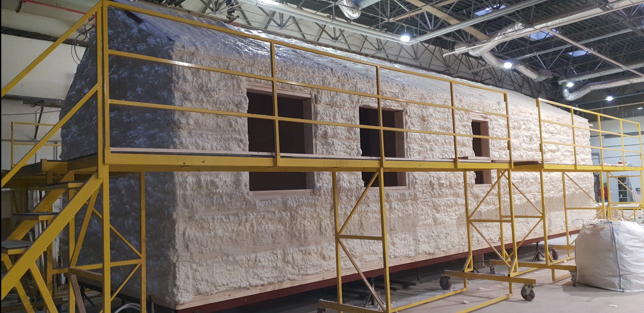 Dom modułowy w trakcie izolacji pianką poliuretanową, widoczny z rusztowania, z otworami okiennymi.