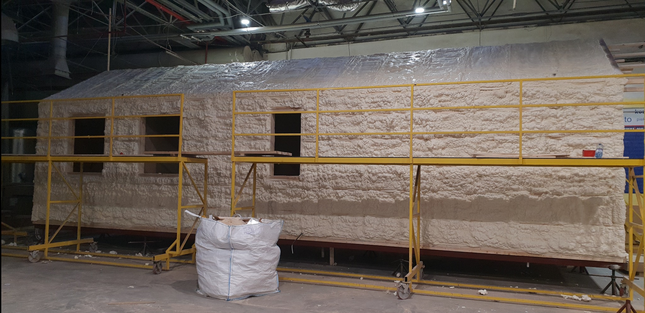 Dom w trakcie izolacji pianką, widoczny z bliska na rusztowaniu w hali, z workiem materiałów budowlanych obok.
