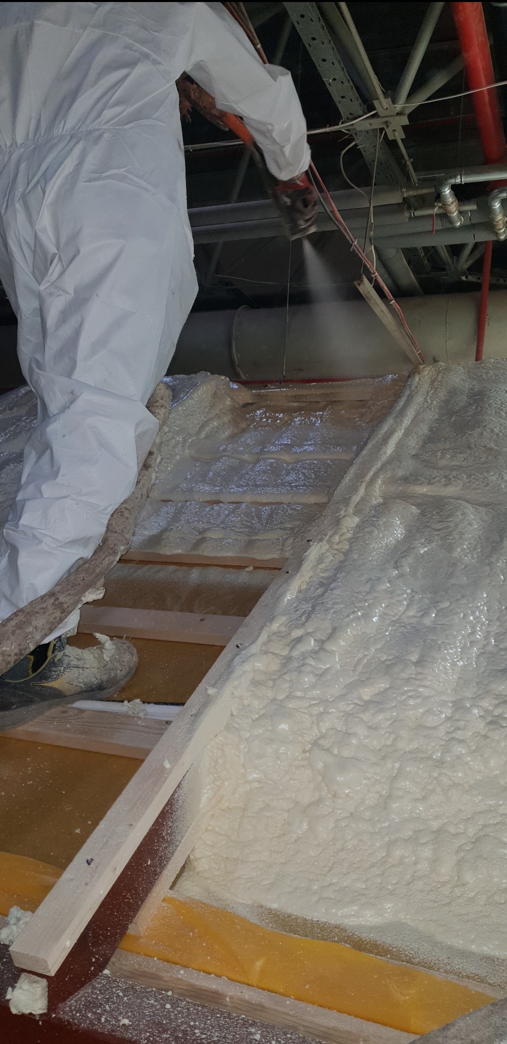 Pracownik w kombinezonie ochronnym natryskuje piankę izolacyjną na konstrukcję poddasza, widoczne drewniane elementy i instalacje pod sufitem.