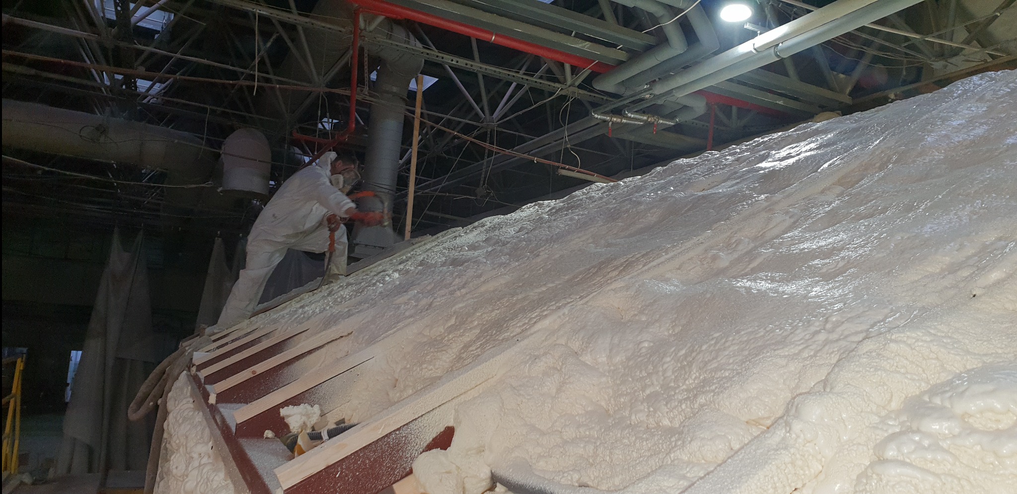 Pracownik w kombinezonie ochronnym natryskuje pianę izolacyjną na konstrukcję dachu w hali przemysłowej, widoczne metalowe elementy konstrukcyjne dachu i instalacje pod sufitem.