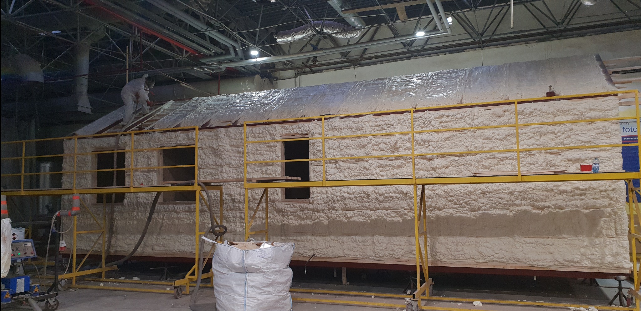 Prace izolacyjne pianką PUR w hali: pracownik w kombinezonie aplikuje pianę na dach konstrukcji szkieletowej, ściany pokryte grubą warstwą pianki, widoczne rusztowania i worek z materiałem.